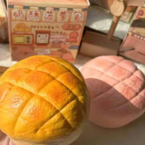 May include: Close-up of two round, textured pastries. One is golden yellow with a diamond pattern, the other is pink. A box with Asian text and illustrations is in the background. The pastries appear to be a type of sweet bread.