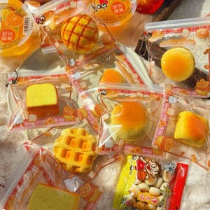 Puede incluir: Se muestra una variedad de productos horneados empaquetados, incluyendo un gofre, rebanadas de pastel y bollos redondos. Los paquetes transparentes tienen un diseño rojo y blanco con el texto "MOOZY BREAD". También hay vasos de gelatina con sabor a naranja y una bolsa de snacks.