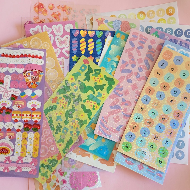 May include: Assortment of colorful sticker sheets with various designs, including flowers, swirls, letters, numbers, and cartoon characters. The stickers are on a pink background.