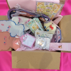 May include: A brown cardboard box filled with various items, including a pink and white plush toy, a sticker sheet, a small notebook, and a keychain. The box is decorated with a pink and white tissue paper and has the words "So much cuteness" written on the bottom.