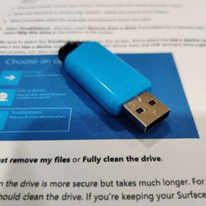 May include: A blue USB flash drive with a silver connector. The drive is on a blue surface with text that reads "Choose an option", "Use a device", "Troubleshoot", "Just remove my files or Fully clean the drive", "The drive is more secure but takes much longer. For", "should clean the drive. If you're keeping your Surface".