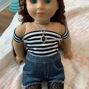 K&ouml;nnte beinhalten: Eine Puppe mit langen braunen Haaren, die ein schwarz-wei&szlig; gestreiftes Shirt mit einem braunen Tanktop darunter, blaue Jeans-Shorts und schwarze Netzstrumpfhosen tr&auml;gt. Die Puppe tr&auml;gt eine silberne Halskette mit einem kleinen ovalen Anh&auml;nger.