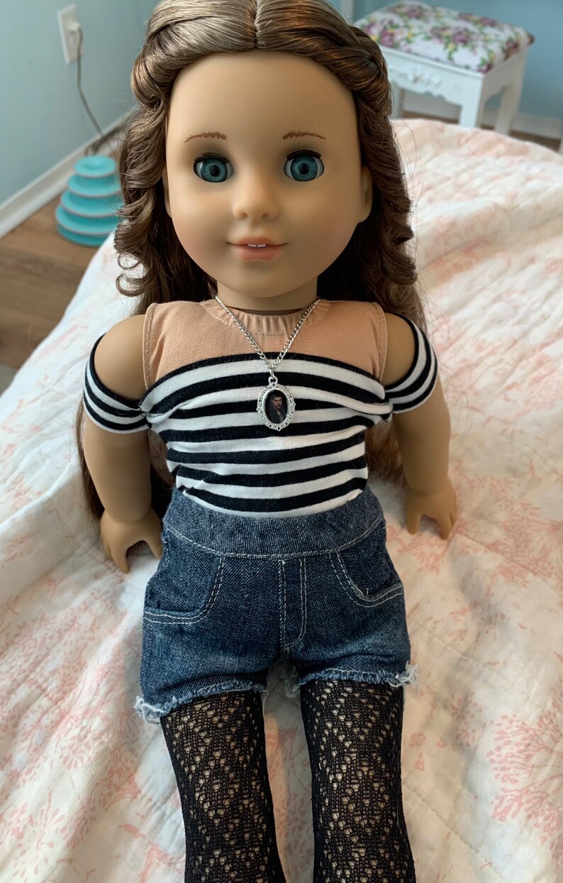 K&ouml;nnte beinhalten: Eine Puppe mit langen braunen Haaren, die ein schwarz-wei&szlig; gestreiftes Shirt mit einem braunen Tanktop darunter, blaue Jeans-Shorts und schwarze Netzstrumpfhosen tr&auml;gt. Die Puppe tr&auml;gt eine silberne Halskette mit einem kleinen ovalen Anh&auml;nger.