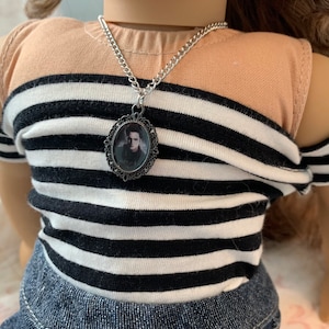 K&ouml;nnte beinhalten: Eine silberne Kettenkette mit einem schwarzen und wei&szlig;en ovalen Anh&auml;nger, der ein Portr&auml;t eines Mannes zeigt. Die Kette wird von einer Puppe mit braunen Haaren und einem schwarz-wei&szlig; gestreiften Shirt getragen.