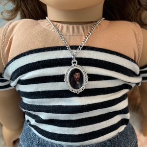 K&ouml;nnte beinhalten: Eine silberne Kettenkette mit einem silbernen ovalen Anh&auml;nger mit einem Schwarzwei&szlig;foto eines Mannes. Die Kette wird von einer Puppe mit braunen Haaren und einem gestreiften Shirt getragen.