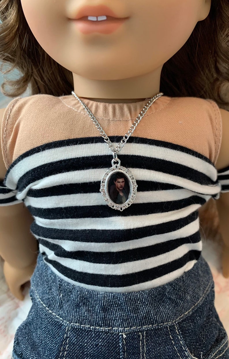 K&ouml;nnte beinhalten: Eine silberne Kettenkette mit einem silbernen ovalen Anh&auml;nger mit einem Schwarzwei&szlig;foto eines Mannes. Die Kette wird von einer Puppe mit braunen Haaren und einem gestreiften Shirt getragen.
