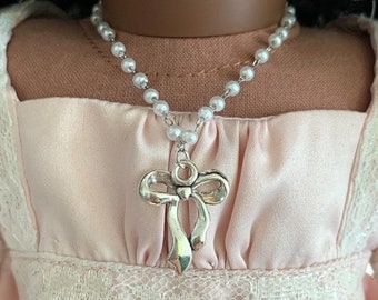 May include: A silver necklace with a pearl chain and a bow pendant. The necklace is worn by a doll with dark hair and a pink dress.