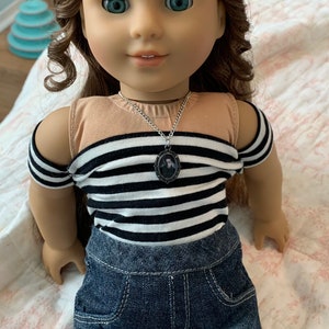 K&ouml;nnte beinhalten: Eine Puppe mit langen braunen Haaren, die ein schwarz-wei&szlig; gestreiftes Shirt, Jeans-Shorts und schwarze Netzstr&uuml;mpfe tr&auml;gt. Die Puppe tr&auml;gt eine silberne Kettenkette mit einem kleinen ovalen Anh&auml;nger.