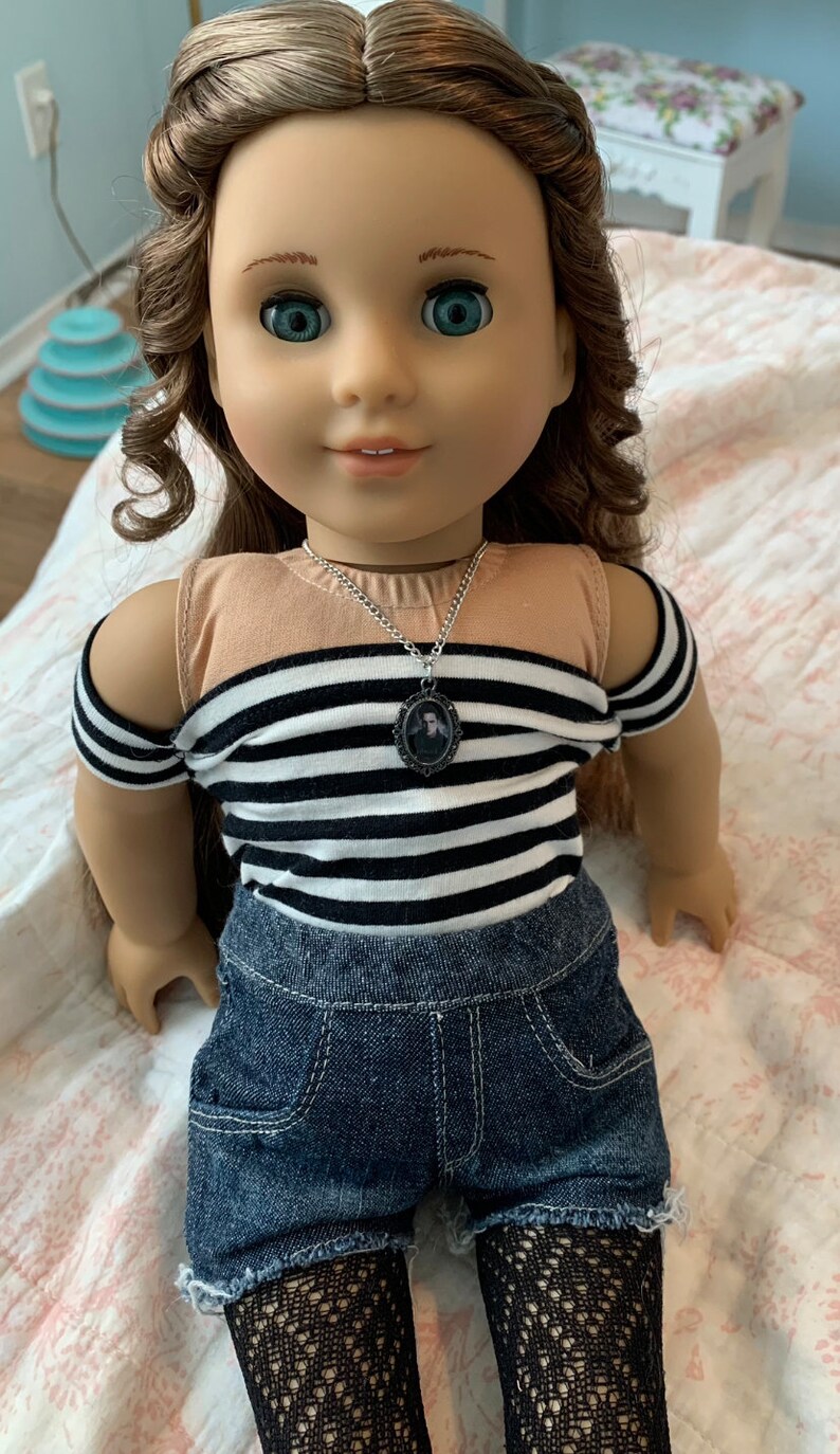 K&ouml;nnte beinhalten: Eine Puppe mit langen braunen Haaren, die ein schwarz-wei&szlig; gestreiftes Shirt, Jeans-Shorts und schwarze Netzstr&uuml;mpfe tr&auml;gt. Die Puppe tr&auml;gt eine silberne Kettenkette mit einem kleinen ovalen Anh&auml;nger.