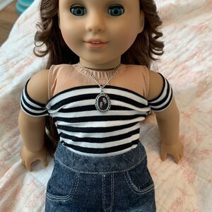 K&ouml;nnte beinhalten: Eine Puppe tr&auml;gt ein schwarz-wei&szlig; gestreiftes Shirt mit einer silbernen Halskette mit einem Foto von zwei Personen. Die Puppe tr&auml;gt au&szlig;erdem Jeans-Shorts und schwarze Netzstr&uuml;mpfe.