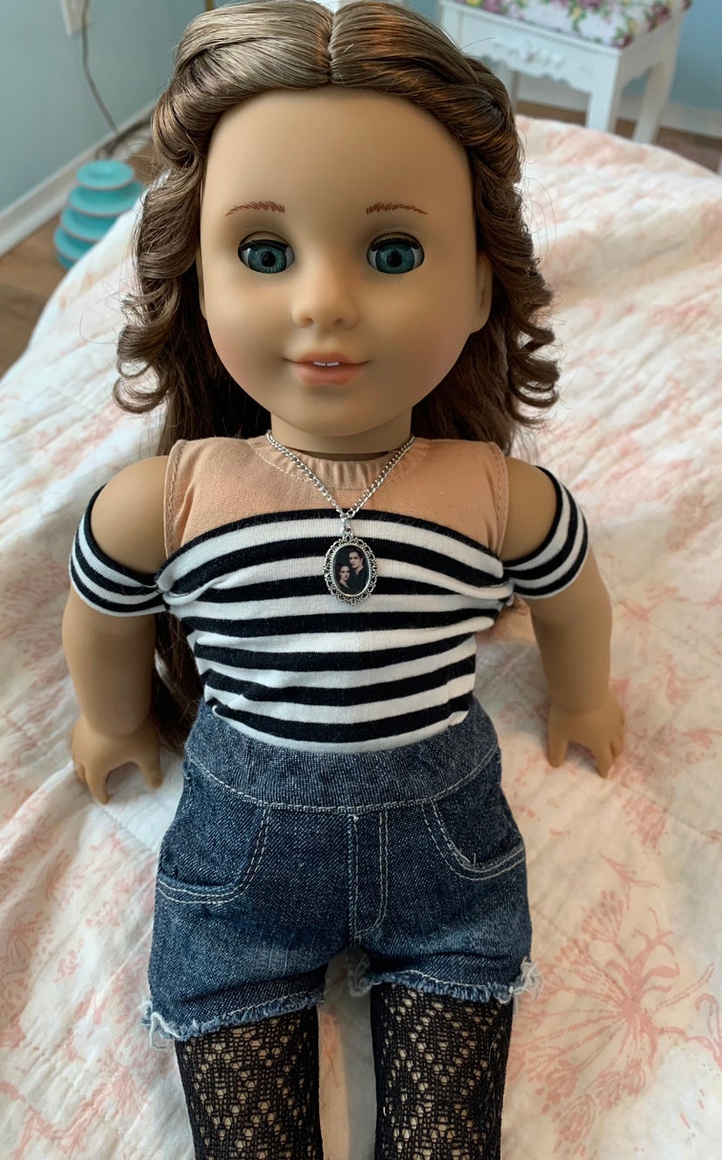 K&ouml;nnte beinhalten: Eine Puppe tr&auml;gt ein schwarz-wei&szlig; gestreiftes Shirt mit einer silbernen Halskette mit einem Foto von zwei Personen. Die Puppe tr&auml;gt au&szlig;erdem Jeans-Shorts und schwarze Netzstr&uuml;mpfe.