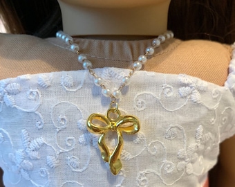 May include: A gold-colored bow pendant necklace with a pearl beaded chain. The necklace is worn on a doll dressed in a white embroidered dress with a pink ribbon.