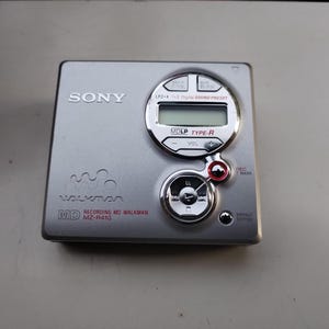 Könnte beinhalten: Ein silberner Sony MD Walkman MZ-R410. Das Gerät hat ein kreisförmiges Display und ein Bedienfeld mit Tasten für Lautstärke, Menü und Aufnahme. Der Text "SONY" und "WALKMAN" sind auf dem Gerät sichtbar.
