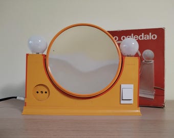 May include: A vintage orange vanity mirror with a round mirror and two white globe lights. The mirror has an orange frame and a white on/off switch. A box with the text "o oaledalo" is in the background.