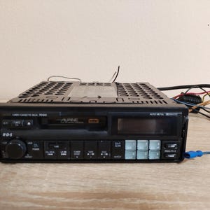 Peut inclure: Autoradio Alpine noir avec lecteur de cassettes et RDS. L'autoradio dispose d'un écran numérique et de plusieurs boutons pour le réglage et le contrôle audio. Des fils et des connecteurs sortent de l'arrière.