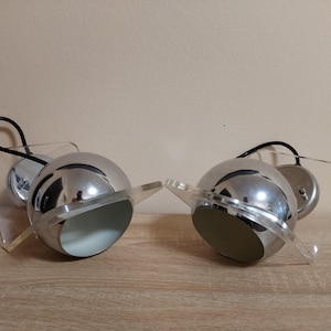 Op de afbeelding: Twee verchroomde, bolvormige wandkandelaars met heldere acrylaccenten. Elke kandelaar heeft een zwart snoer en een ronde basis. De wandkandelaars staan op een houten oppervlak tegen een neutrale achtergrond.