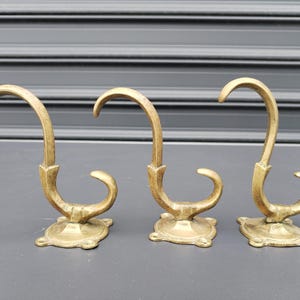 May include: Three antique brass wall hooks. Each hook has a curved design with a decorative base. The hooks are made of a gold-coloured metal and are arranged in a row against a grey background.