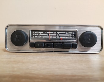May include: A vintage car radio with a chrome faceplate and black knobs. The radio features a frequency dial with numbers and letters, and several black buttons. The radio is designed to fit into a car dashboard.