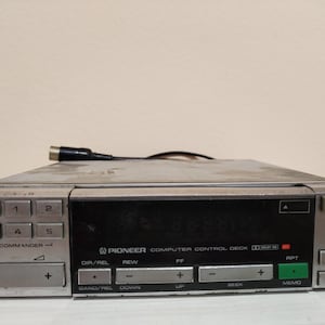 May include: A vintage Pioneer Computer Control Deck with a silver and black finish. The deck features multiple buttons, including "SDK", "1", "2", "3", "4", "5", "EJECT", and "MEMO". A cable with a connector is plugged into the device.