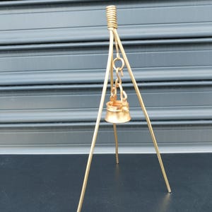 以下が含まれることがあります： 中央に小さなベルが吊り下げられた、金色の金属製の装飾的な三脚。三脚には3本の脚とコイル状のトップが付いています。ベルも金色で丸みを帯びた形をしています。