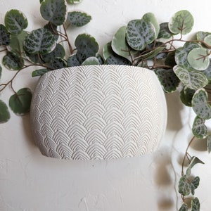 May include: A white, textured, oval-shaped planter with a repeating fan-like pattern. Green and white trailing plant stems and leaves cascade from the top of the planter, creating a natural, decorative display. The planter is mounted on a white wall.