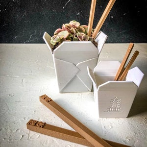 May include: Two white, origami-style planters shaped like takeaway boxes, one filled with a small plant and wooden chopsticks. A second, smaller planter is visible, along with two pairs of brown chopsticks.