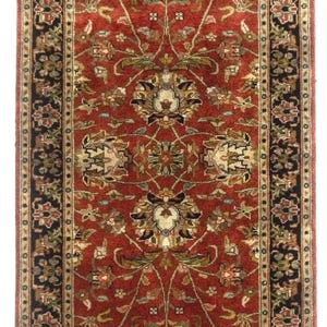May include: A long, rectangular runner rug featuring a deep red field decorated with symmetrical floral motifs in shades of green, cream, and black. The rug is framed by a wide, dark border with intricate repeating floral patterns and fringe.