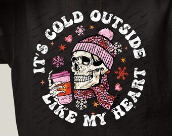 May include: Black sweatshirt with a white graphic print of a skull wearing a pink hat and scarf holding a cup of coffee. The text around the skull reads "It's cold outside like my heart".