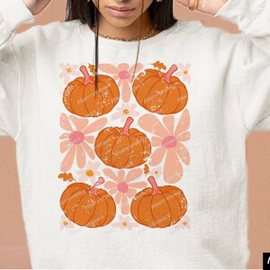 May include: A white sweatshirt with a graphic design of orange pumpkins and pink flowers. The pumpkins have a distressed look and are arranged in a grid pattern.