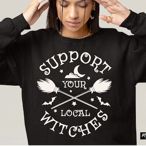 Puede incluir: Una sudadera negra con un diseño gráfico blanco que dice "Support Your Local Witches". El gráfico incluye una luna creciente, estrellas y dos escobas cruzadas.