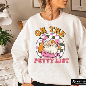 May include: White sweatshirt with a graphic of Santa Claus in a pink outfit holding a bag of gifts. The graphic is surrounded by a checkered pattern and the text "On the Petty List".