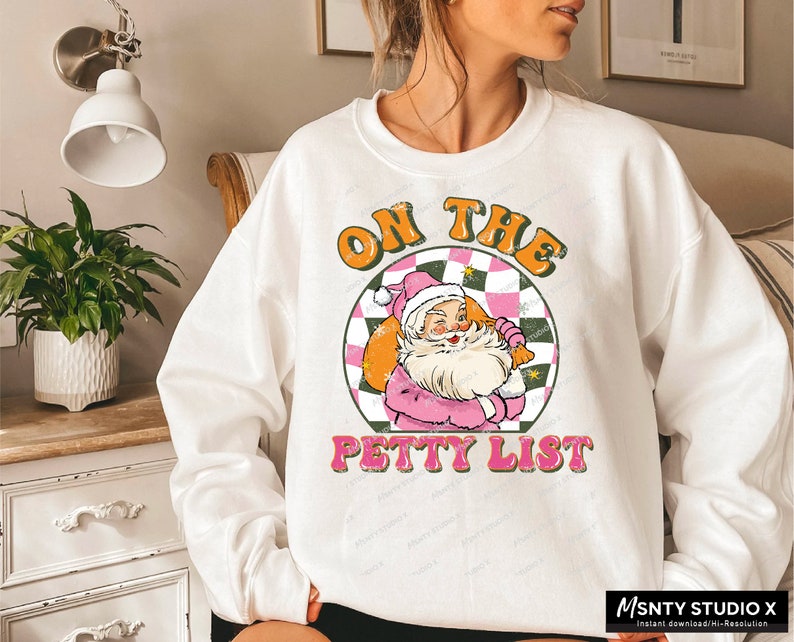 May include: White sweatshirt with a graphic of Santa Claus in a pink outfit holding a bag of gifts. The graphic is surrounded by a checkered pattern and the text "On the Petty List".