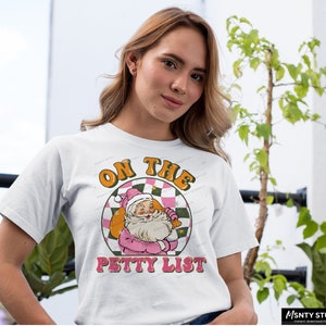 May include: White t-shirt with a pink and green graphic of Santa Claus with the text "On the Petty List".