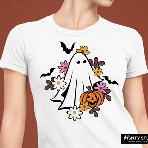 Pu&ograve; includere: Un fantasma bianco con un viso amichevole circondato da fiori rosa, arancioni e marroni e una zucca di Halloween. Il design &egrave; su una maglietta bianca.