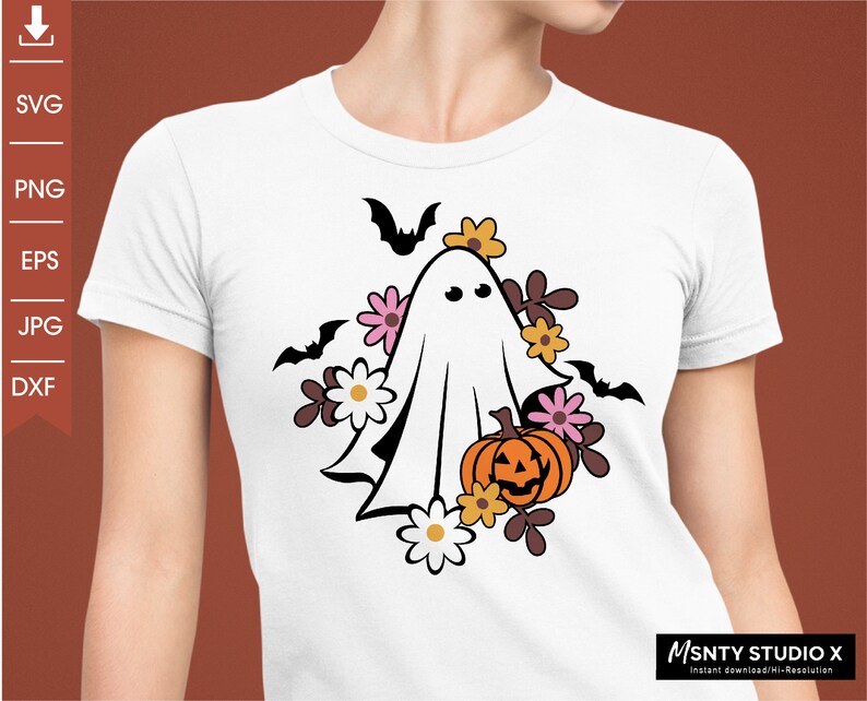 Pu&ograve; includere: Un fantasma bianco con un viso amichevole circondato da fiori rosa, arancioni e marroni e una zucca di Halloween. Il design &egrave; su una maglietta bianca.