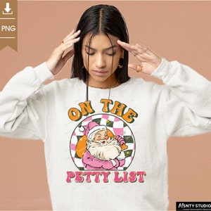 May include: A white sweatshirt with a pink and green graphic design featuring a cartoon Santa Claus holding a bag of gifts. The graphic text reads "On the Petty List".