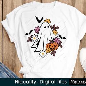 Pu&ograve; includere: Una maglietta bianca con un fantasma, una zucca e fiori in stile retr&ograve;. Il fantasma ha un viso amichevole ed &egrave; circondato da fiori rosa, gialli e arancioni. La zucca ha un viso di zucca.
