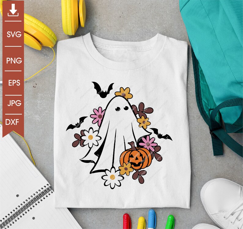 Pu&ograve; includere: Un fantasma bianco con un viso amichevole che tiene una zucca intagliata a forma di viso. Il fantasma &egrave; circondato da foglie marroni e fiori rosa e bianchi. Il design &egrave; su una maglietta bianca.