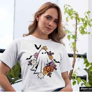 Pu&ograve; includere: Una maglietta bianca con una grafica di un fantasma che indossa una corona di fiori e tiene una zucca. Il fantasma &egrave; circondato da fiori e pipistrelli.