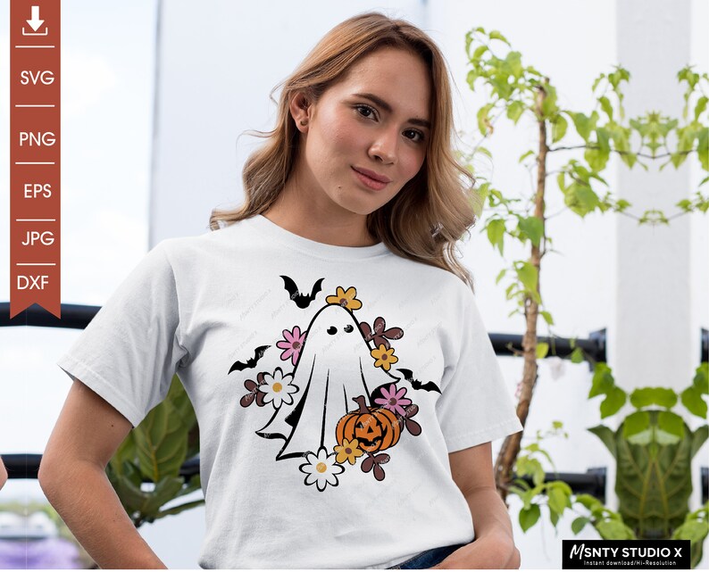 Pu&ograve; includere: Una maglietta bianca con una grafica di un fantasma che indossa una corona di fiori e tiene una zucca. Il fantasma &egrave; circondato da fiori e pipistrelli.