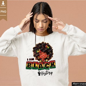 K&ouml;nnte beinhalten: Ein wei&szlig;er Pullover mit einem Grafikdesign, das ein schwarzes Cartoon-M&auml;dchen mit einer rot-gelb-gr&uuml;nen Afrofrisur zeigt. Der Text "I AM BLACK History" ist in schwarzen Buchstaben auf dem Pullover gedruckt.