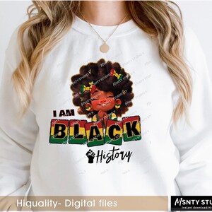 K&ouml;nnte beinhalten: Ein wei&szlig;es Sweatshirt mit einem Grafikdesign, das ein schwarzes Cartoon-M&auml;dchen mit einem rot-gelb-gr&uuml;nen Stirnband zeigt. Der Text "I AM BLACK HISTORY" ist in schwarzen Buchstaben auf einem rot-gelb-gr&uuml;nen Hintergrund gedruckt.