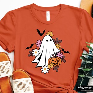 Pu&ograve; includere: Una maglietta arancione con un fantasma bianco che tiene una zucca di Halloween circondata da fiori e pipistrelli.