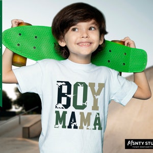 Pu&ograve; includere: Una maglietta bianca con un design grafico verde, nero e bianco che dice "BOY MAMA" con silhouette di dinosauri nelle lettere. Il design &egrave; ispirato al camouflage. Un bambino piccolo indossa la maglietta e tiene uno skateboard verde.