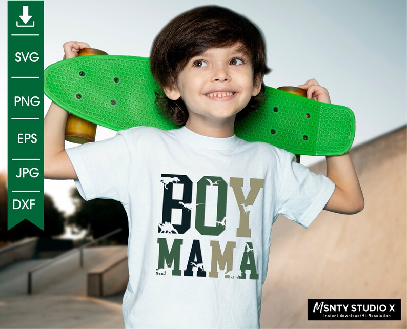 Pu&ograve; includere: Una maglietta bianca con un design grafico verde, nero e bianco che dice "BOY MAMA" con silhouette di dinosauri nelle lettere. Il design &egrave; ispirato al camouflage. Un bambino piccolo indossa la maglietta e tiene uno skateboard verde.