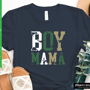 Pu&ograve; includere: Una maglietta blu navy con il testo "BOY MAMA" in lettere bianche, verdi e dorate. Le lettere sono circondate da silhouette di dinosauri.