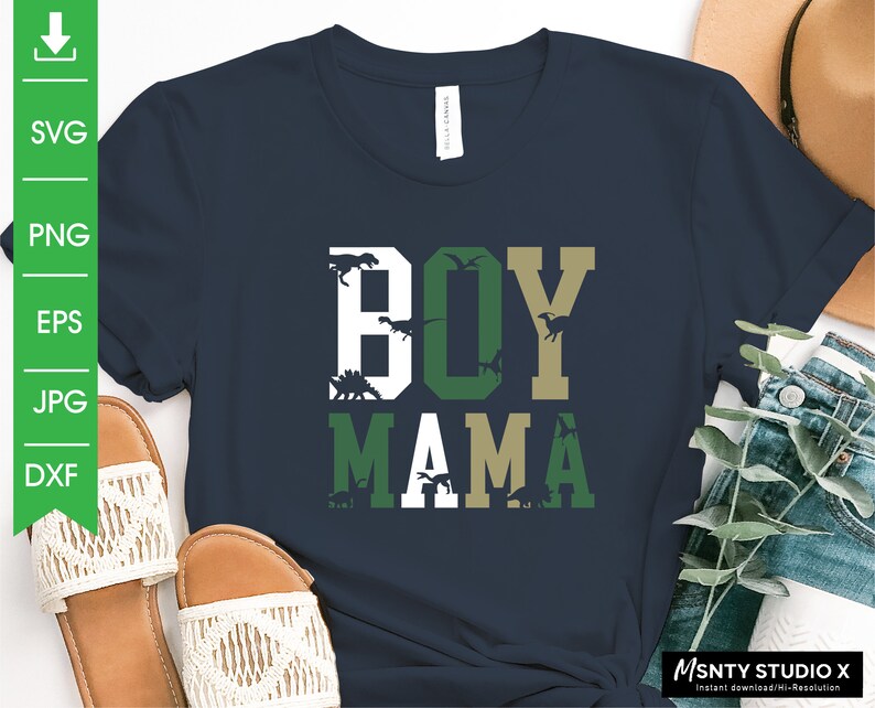 Pu&ograve; includere: Una maglietta blu navy con il testo "BOY MAMA" in lettere bianche, verdi e dorate. Le lettere sono circondate da silhouette di dinosauri.