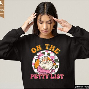 May include: Black sweatshirt with a pink and green graphic of Santa Claus with the text "On the Petty List".