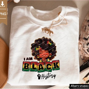 K&ouml;nnte beinhalten: Ein wei&szlig;es T-Shirt mit einem Grafikdesign, das ein Cartoon-M&auml;dchen mit brauner Haut und lockigem Haar zeigt. Das M&auml;dchen tr&auml;gt ein rot-gelb-gr&uuml;nes Stirnband und h&auml;lt ihre H&auml;nde in einer Gebetsposition zusammen. Der Text "I AM BLACK History" ist in rot-gelb-gr&uuml;nen Buchstaben unter dem M&auml;dchen gedruckt.