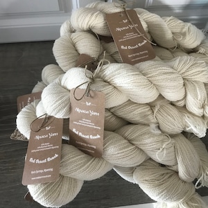 May include: Several skeins of cream-coloured alpaca yarn with attached tags from Red Desert Ranch. The tags are brown and feature the text "Alpaca Yarn" and "Red Desert Ranch". The yarn is displayed on a wooden surface. A decorative wreath hangs in the background.